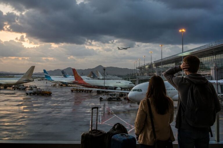 Two travelers with rolling suitcases stand by a terminal window, watching planes on the wet tarmac as a jet climbs into a cloudy sky.
