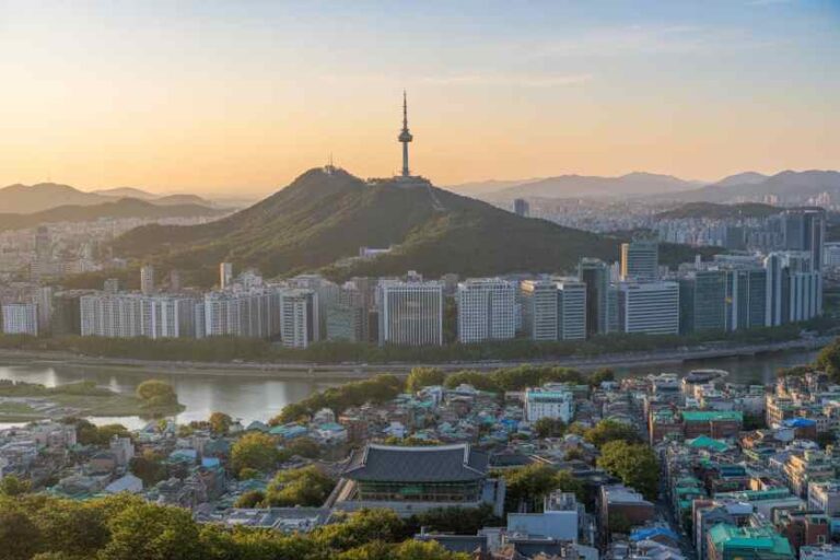 Namsan Mountain with the Seoul Tower atop, overlooking a dense urban skyline at sunset by the river.