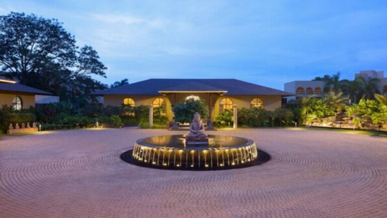 Evening view of a circular fountain with a seated Buddha statue in the center, illuminated in a landscaped courtyard surrounded by plants and arches.