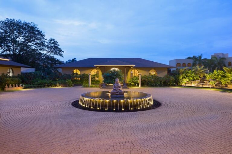 Circular brick courtyard around a illuminated fountain with a meditating statue at the center, night-time garden and one-story building with arched windows in the background.
