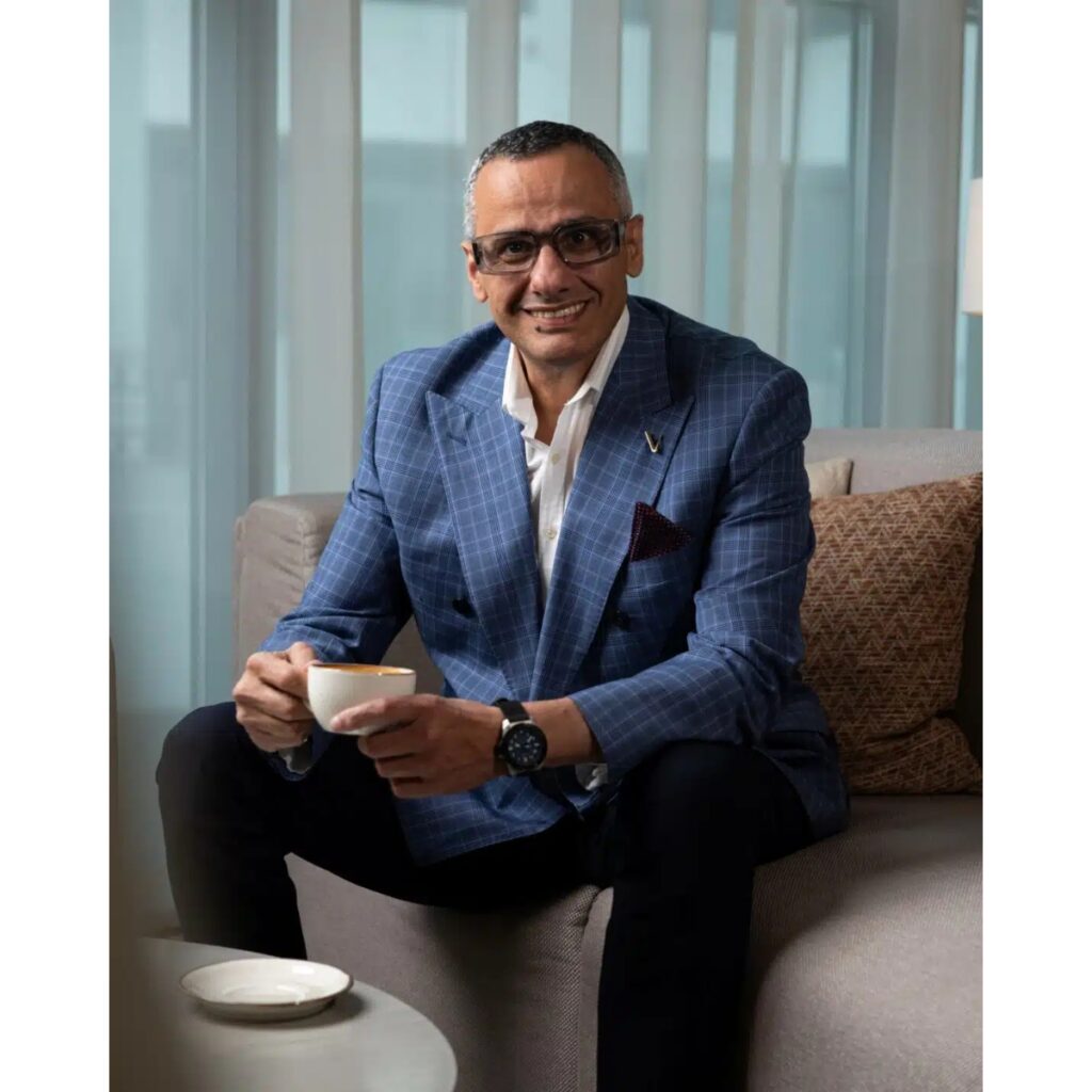 Smiling man in a blue check blazer sits on a sofa, holding a cup of coffee in a bright lounge.