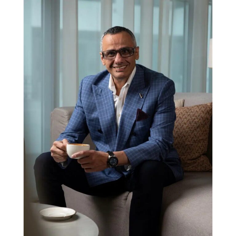 Smiling man in a blue check blazer sits on a sofa, holding a cup of coffee in a bright lounge.