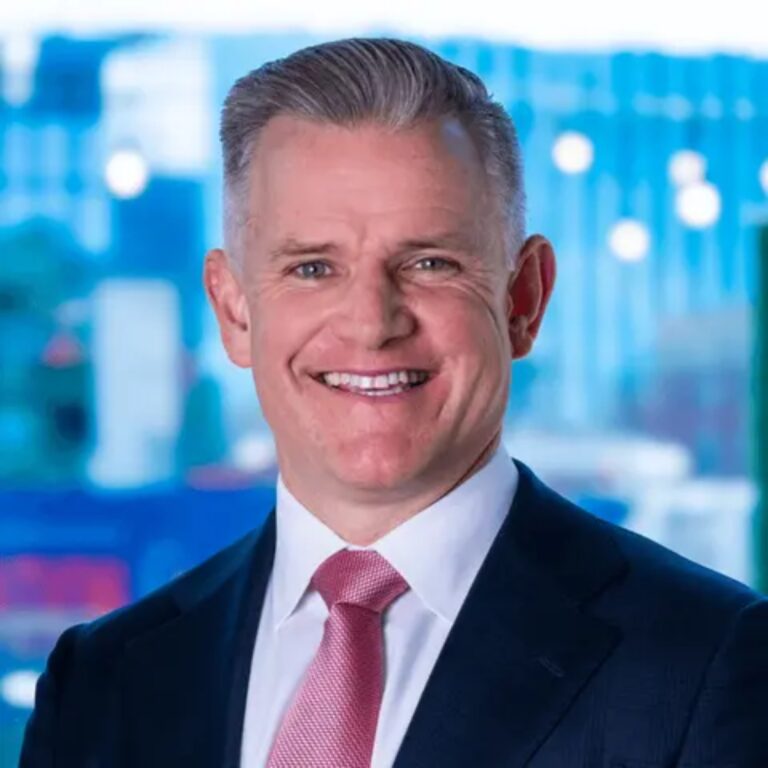 Smiling man in a dark suit and pink tie against a blue, blurred office background.