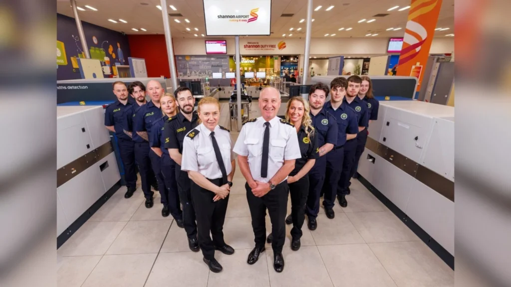 Group of Shannon Airport staff in navy and white uniforms posing for a team photo in a busy security checkpoint area.