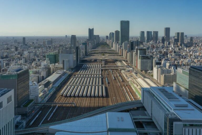 Bird's-eye view of a dense city with multiple parallel railway tracks and long platforms extending toward a distant skyline of tall buildings under a clear blue sky.
