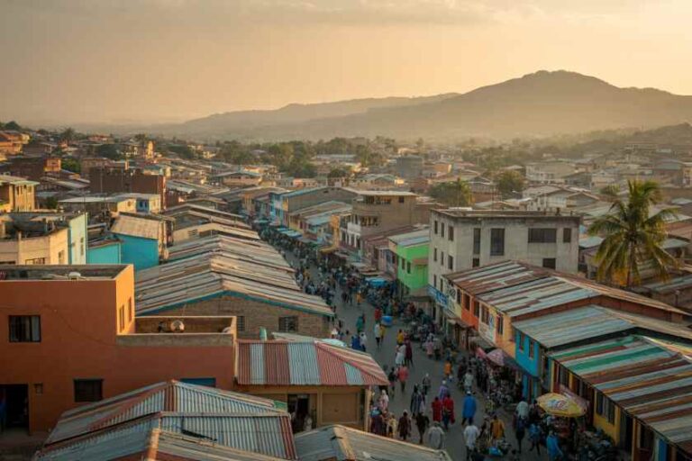 A bustling street market in a town at sunset, with colorful corrugated-roof shops lining a crowded avenue and hills in the distance.
