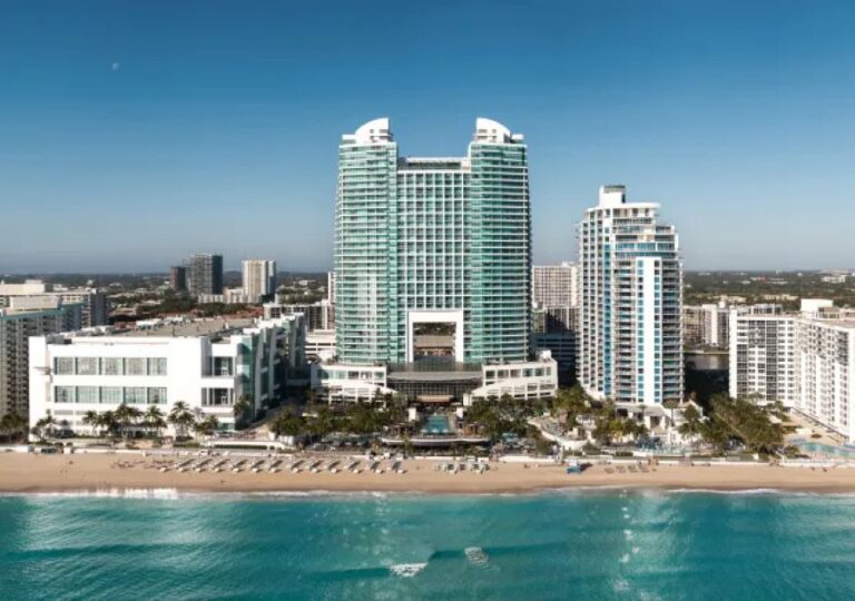 A beachfront hotel complex with two tall green glass towers and a white facade along a sandy shore, turquoise sea in the foreground.