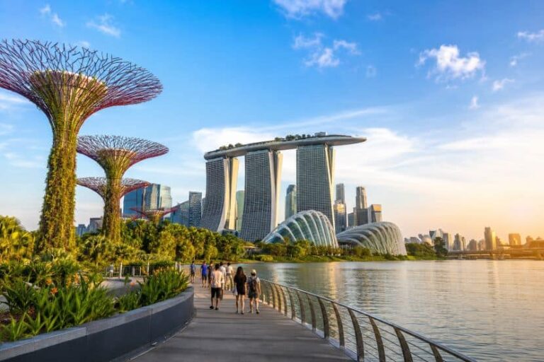 People walk along a waterfront promenade with Supertree Grove and Marina Bay Sands in Singapore under a blue sky.