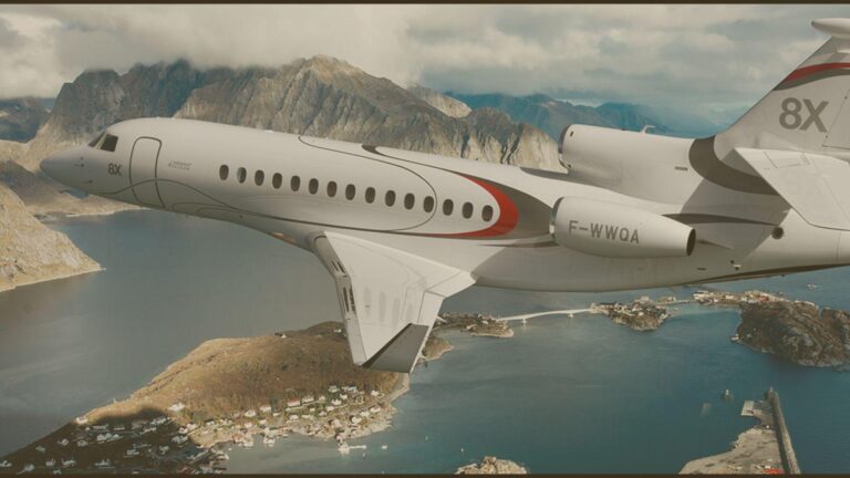 White private jet in flight over a rocky coastline with mountains and a calm blue sea below.