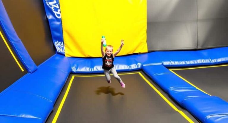 A young child jumps mid-air on a blue trampoline with yellow edge padding in an indoor trampoline park. online park area in background shows bold blue and yellow colors.
