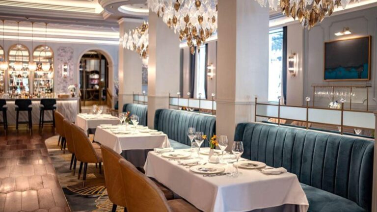 Upscale restaurant dining area with white-tablecloth tables along a blue velvet banquette and warm chandeliers overhead