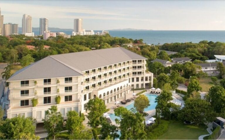 A large beige hotel with a curved façade and multiple balconies, adjacent to a rectangular pool surrounded by lounge chairs and lush trees, with the ocean in the distance and a city skyline.