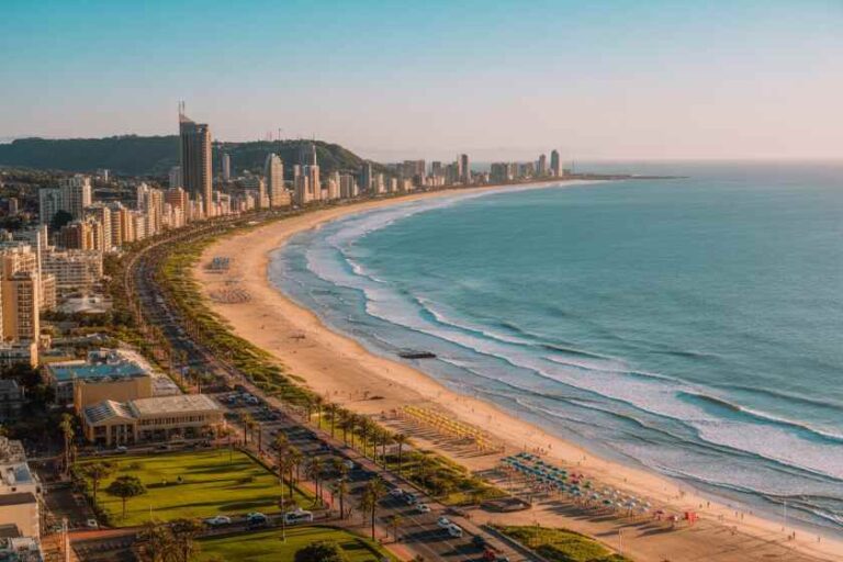 Aerial view of a sunny coastal city with a long sandy beach and high-rise buildings along the shore, turquoise water to the right.