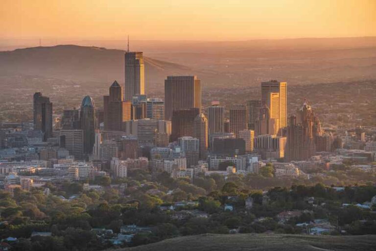 Sunlit city skyline at sunset with tall office towers and a hazy orange sky above a green suburban foreground.