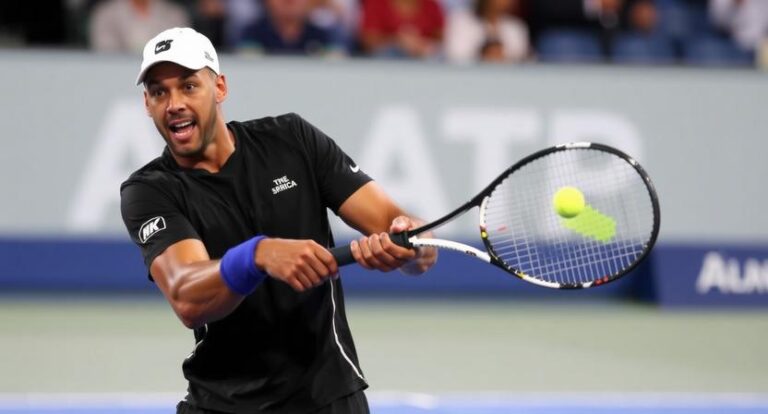 Tennis player in a black shirt and white cap hits a forehand on a court, crowd in the stands behind him.