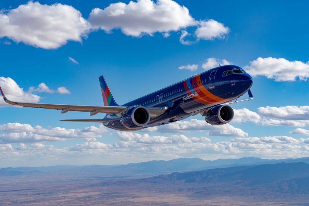 Blue passenger jet with orange and red accents flying over a desert landscape with mountains and scattered clouds in a bright blue sky.