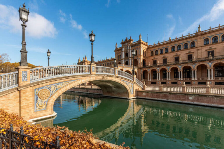 Sunlit view of the Plaza de España in Seville showing an ornate arched bridge over a canal and a grand brick building in the background with blue sky above.