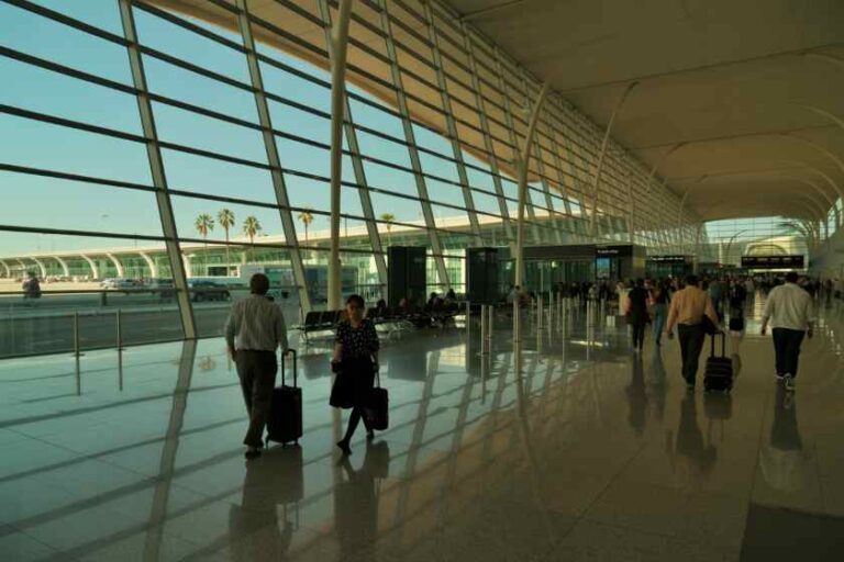 Busy airport terminal concourse with travelers pulling rolling suitcases along a polished floor beside tall glass walls and curved ceiling rails.