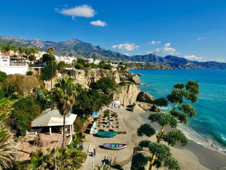 Coastal village perched on cliffs with white buildings, palm trees, and turquoise sea under a blue sky.
