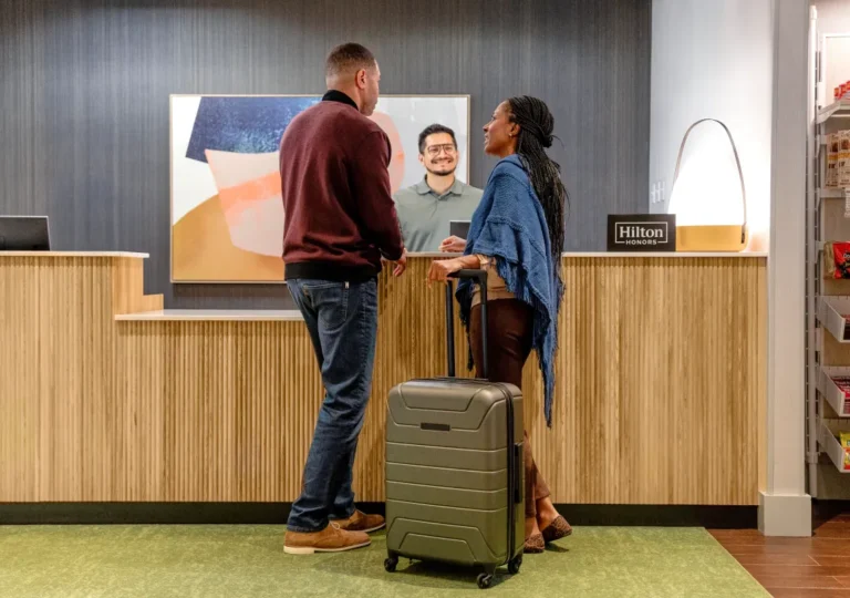 Two guests check in at a hotel reception while a staff member smiles behind the desk, with a green suitcase in the foreground.