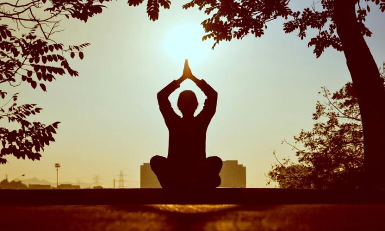 Silhouette of a person in a cross-legged lotus position, hands pressed together overhead, meditating at sunset framed by trees.