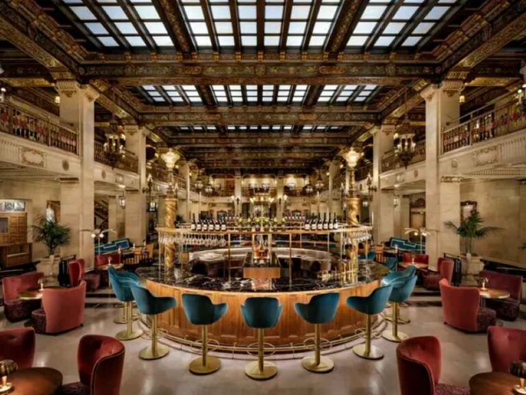 Luxurious hotel bar with a circular dark marble counter surrounded by turquoise bar stools in a grand, ornate hall with balconies and warm lighting