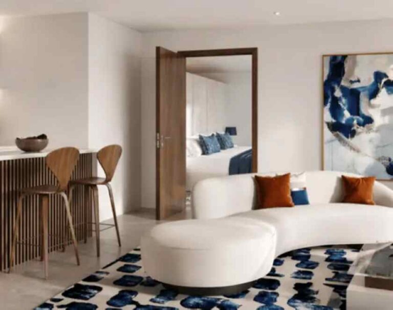 Open-plan living area with a white curved sofa, blue pillows, and a blue-pattern rug; doorway leads to a bright bedroom beyond.