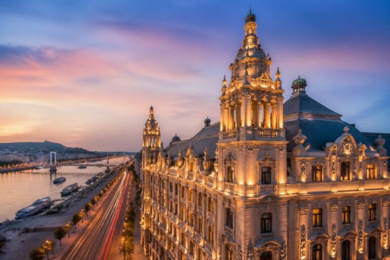 Ornate illuminated historic building along a river at dusk, with a busy street and light trails in the foreground.