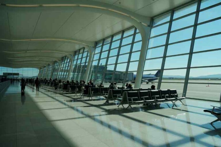 Airport terminal waiting area with rows of seats and large glass walls, a plane visible on the tarmac outside.