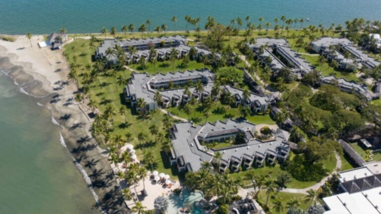 Aerial view of a coastal resort with low-rise buildings, palm trees, green lawns, and a sandy beach along turquoise water.