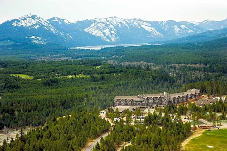 Aerial view of a mountain resort with a large lodge complex surrounded by pine forest and snow‑capped peaks in the distance.