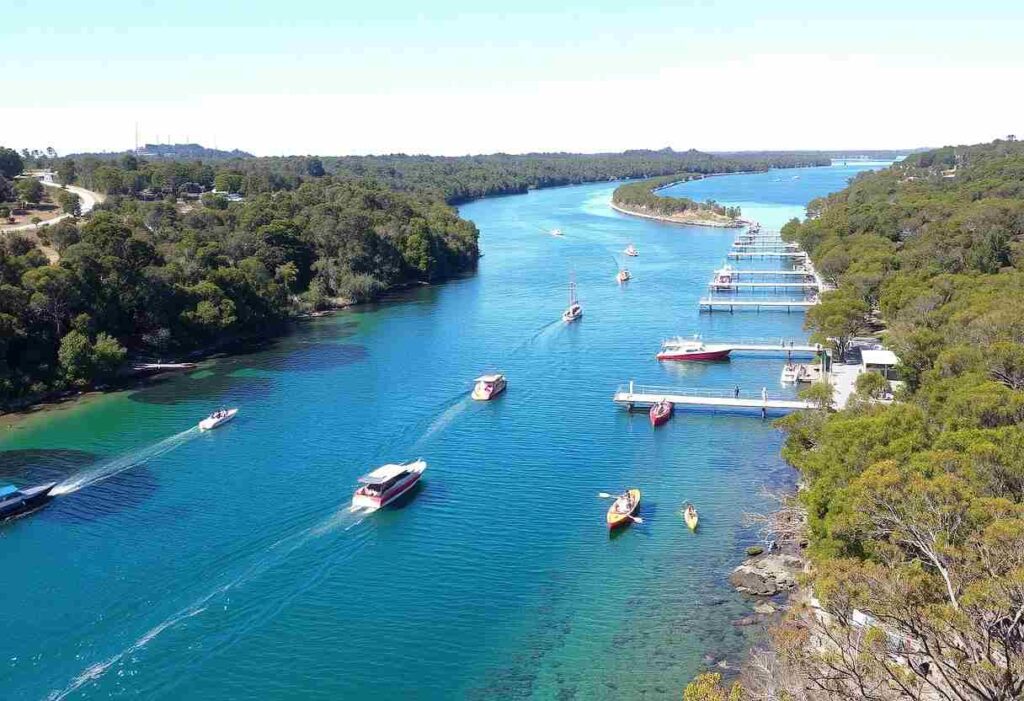 Aerial view of a blue river with several boats cruising, tree-lined banks, and multiple floating docks along the right side.