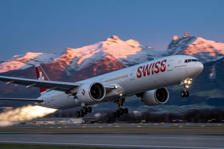 SWISS airplane lifting off from a runway with snow-capped mountains in the background at sunset/sunrise.
