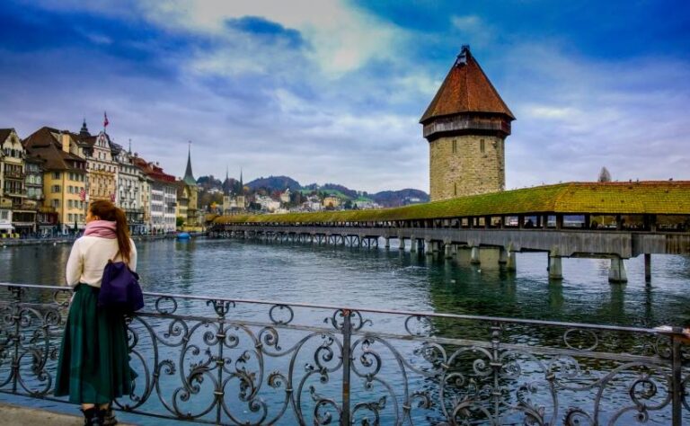 Woman with a purple backpack gazes at Lucerne's Kapellbrücke covered bridge crossing a calm river, ornate railing in the foreground