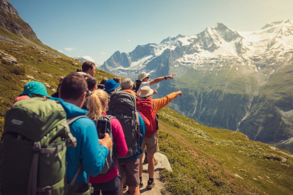 Group of hikers with backpacks on a mountain trail, pointing toward the snow‑capped peaks in the distance.