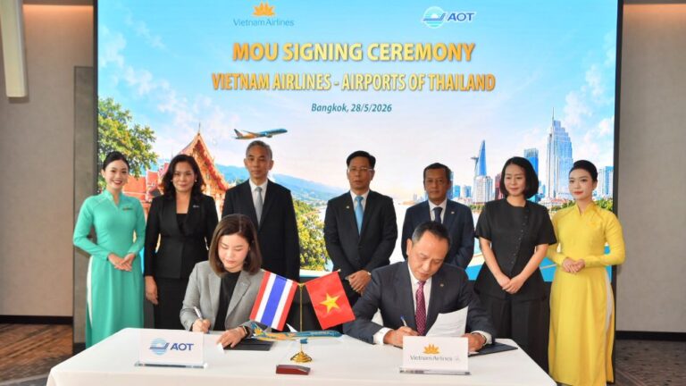 MOU signing between Vietnam Airlines and Airports of Thailand with two officials signing at a table, Thai and Vietnamese flags displayed, group of officials stand behind under a banner in Bangkok.