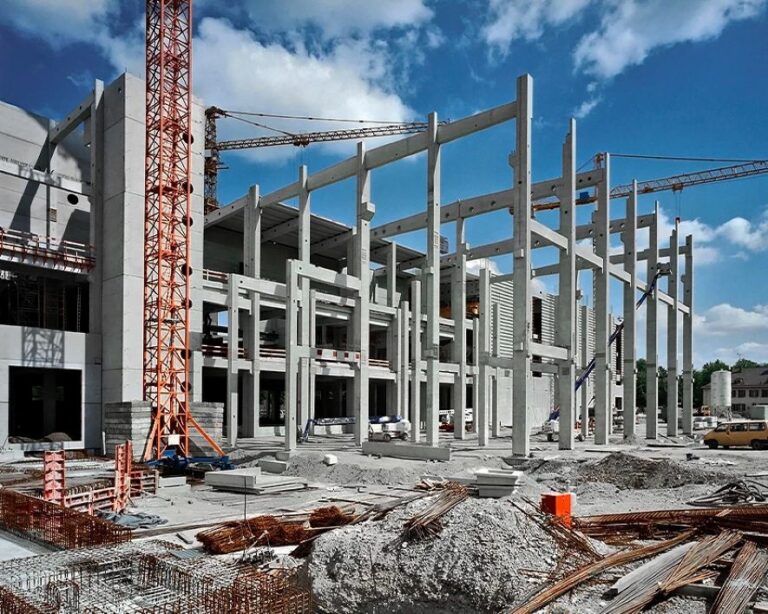 Construction site with a multi-story concrete frame and cranes overhead under clear blue sky.