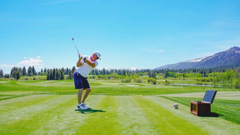 Golfer mid-swing on a sunny green course with mountains in the distance; a laptop on a wooden stand sits to the right.