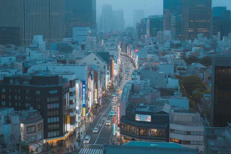 Aerial view of a busy city street lined with tall buildings and bright storefronts, with cars and pedestrians in the avenue far below the high-rises in the distance.