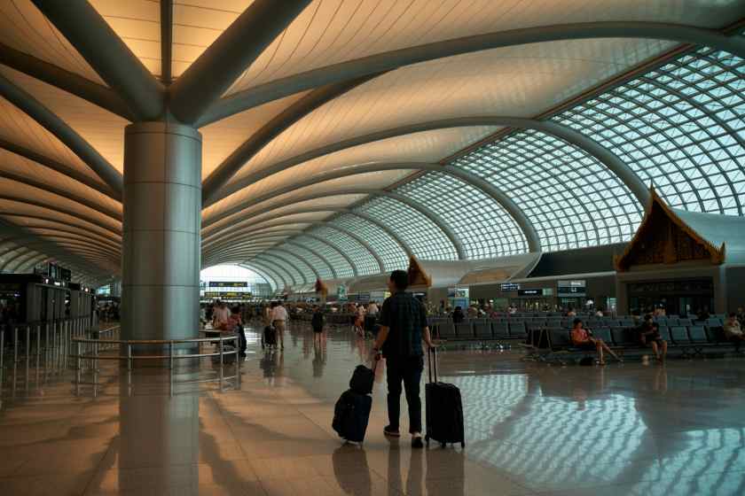 Traveler with two rolling suitcases stands in a bright, spacious airport terminal under a curved glass roof with supporting columns and rows of seats in the background.