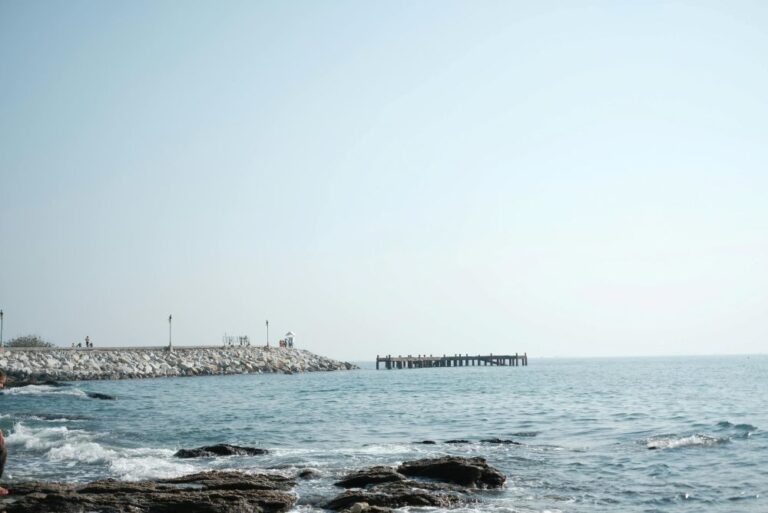 Rocky shoreline with a breakwater extending into the calm sea under a clear blue sky.