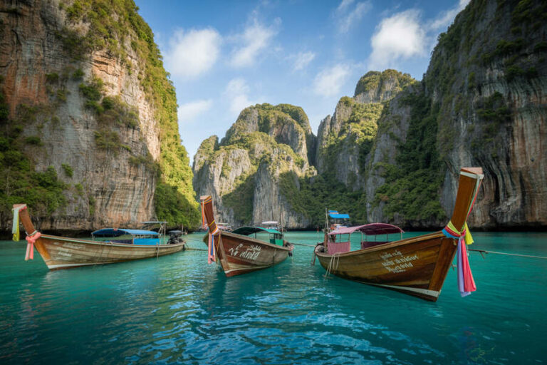 Three traditional Thai long-tail boats anchored in a turquoise lagoon, stone cliffs looming with greenery in the background.
