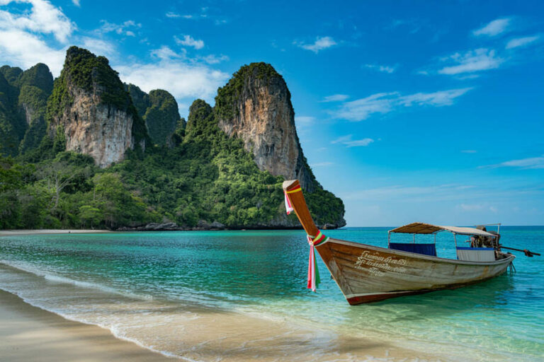 Long-tail boat anchored on a turquoise tropical sea with towering green cliffs and blue sky behind.