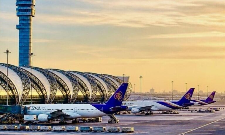 Thai Airways planes parked at a gate at sunrise with the curved glass concourse in the foreground and a tall control tower nearby.