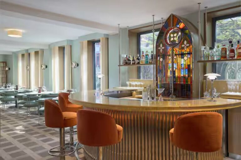 Curved marble bar with orange upholstered stools in a retro-modern lounge; stained-glass arch and liquor shelves behind the counter.