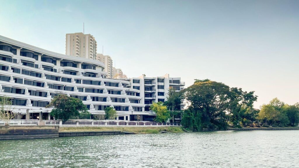 White multi-story residential complex along a river with trees in the foreground and a promenade view.