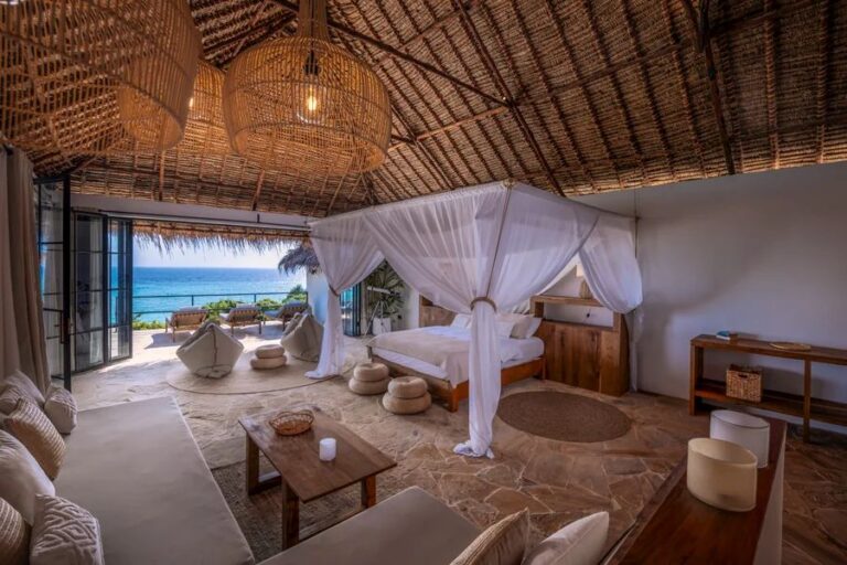 Open-air tropical bedroom with a four-poster bed draped in white fabric, wicker lanterns, and a view of the ocean.