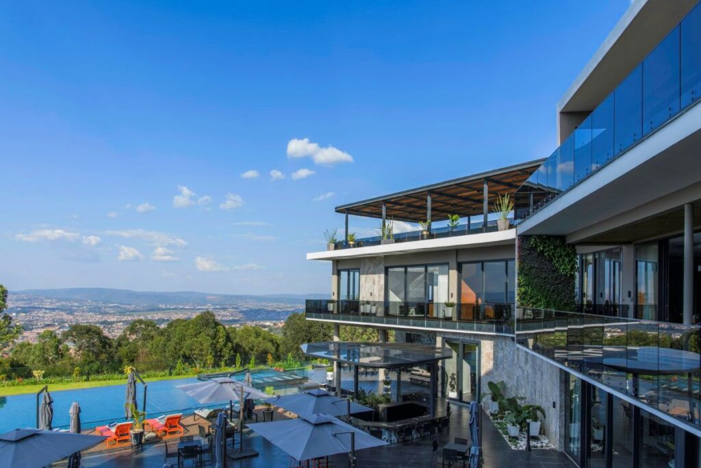 Modern hillside hotel with glass balconies overlooking an outdoor pool and lounge area under a blue sky.