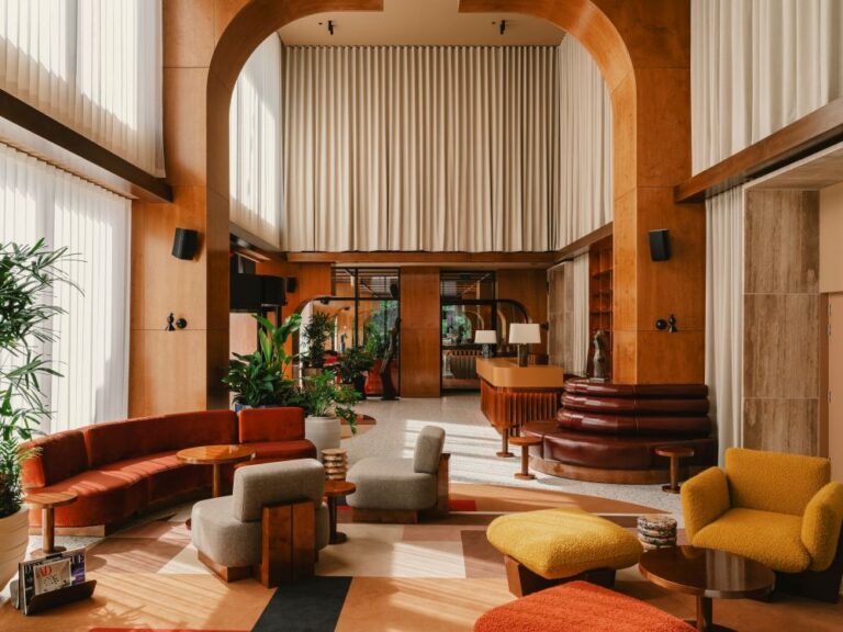 Spacious hotel lobby with curved red sofa, yellow chairs, and round wood tables amid green plants and warm wood panels.