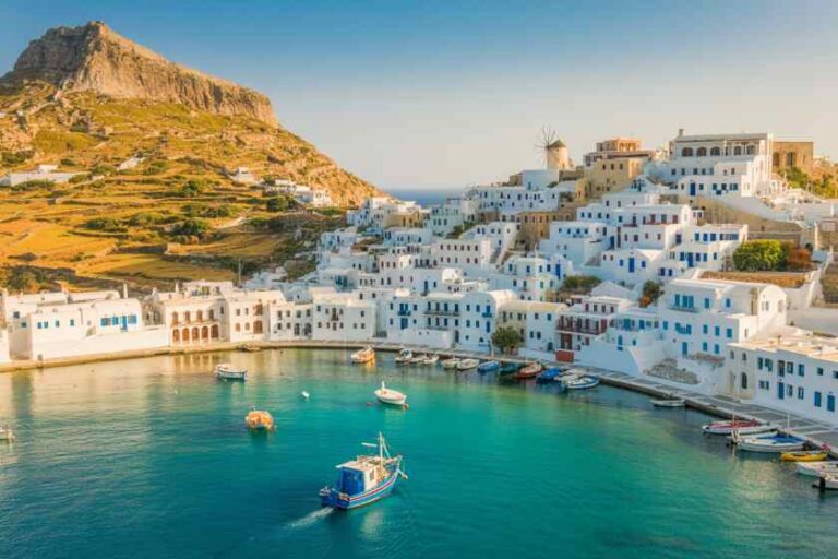 White-washed buildings stacked on a hillside above a turquoise Greek harbor with small boats.